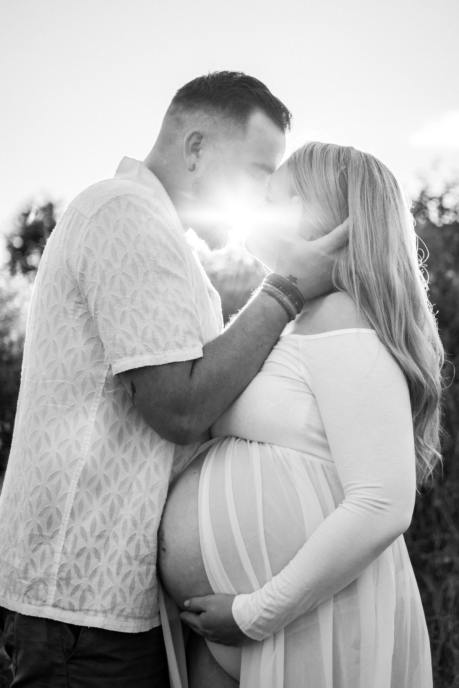 Pregnant couple sharing a tender kiss outdoors, backlit by golden sunlight, woman in white maternity dress cradling her baby bump.