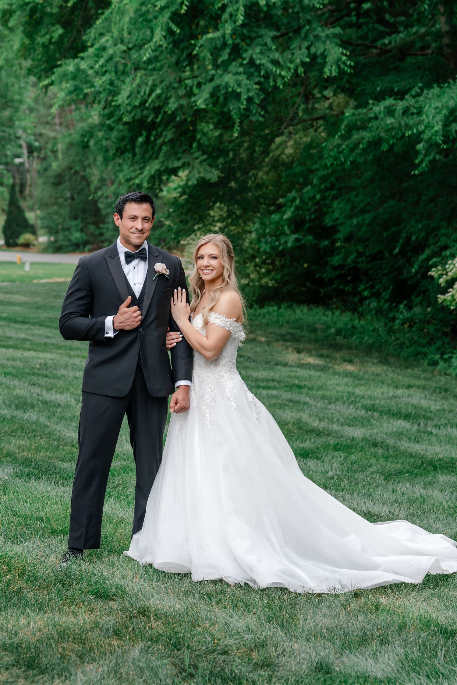 A bride and groom pose together on a lush green lawn, she displays her ring while wearing an off-shoulder gown.