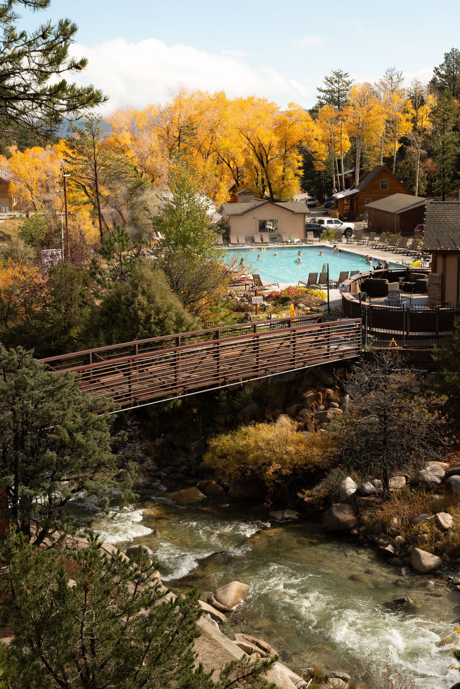 Rustic wooden bridge spans across rushing river with fall foliage and mountain scenery.