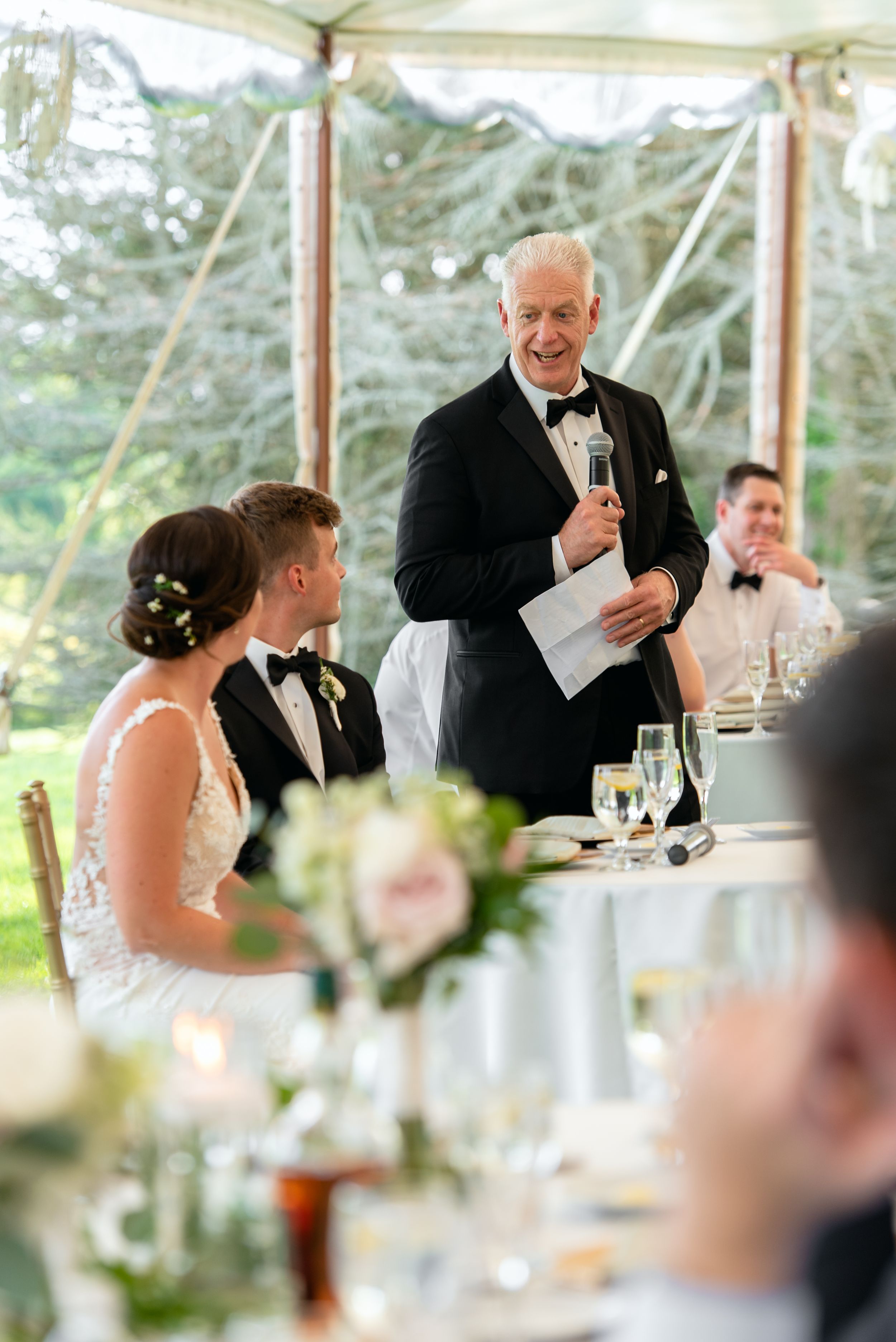 Older man in tuxedo giving wedding speech at elegant outdoor reception while bride and groom listen.
