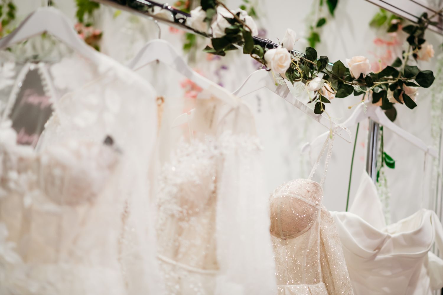 Elegant white lace wedding dresses displayed on hangers adorned with pink roses and greenery in a bridal boutique.