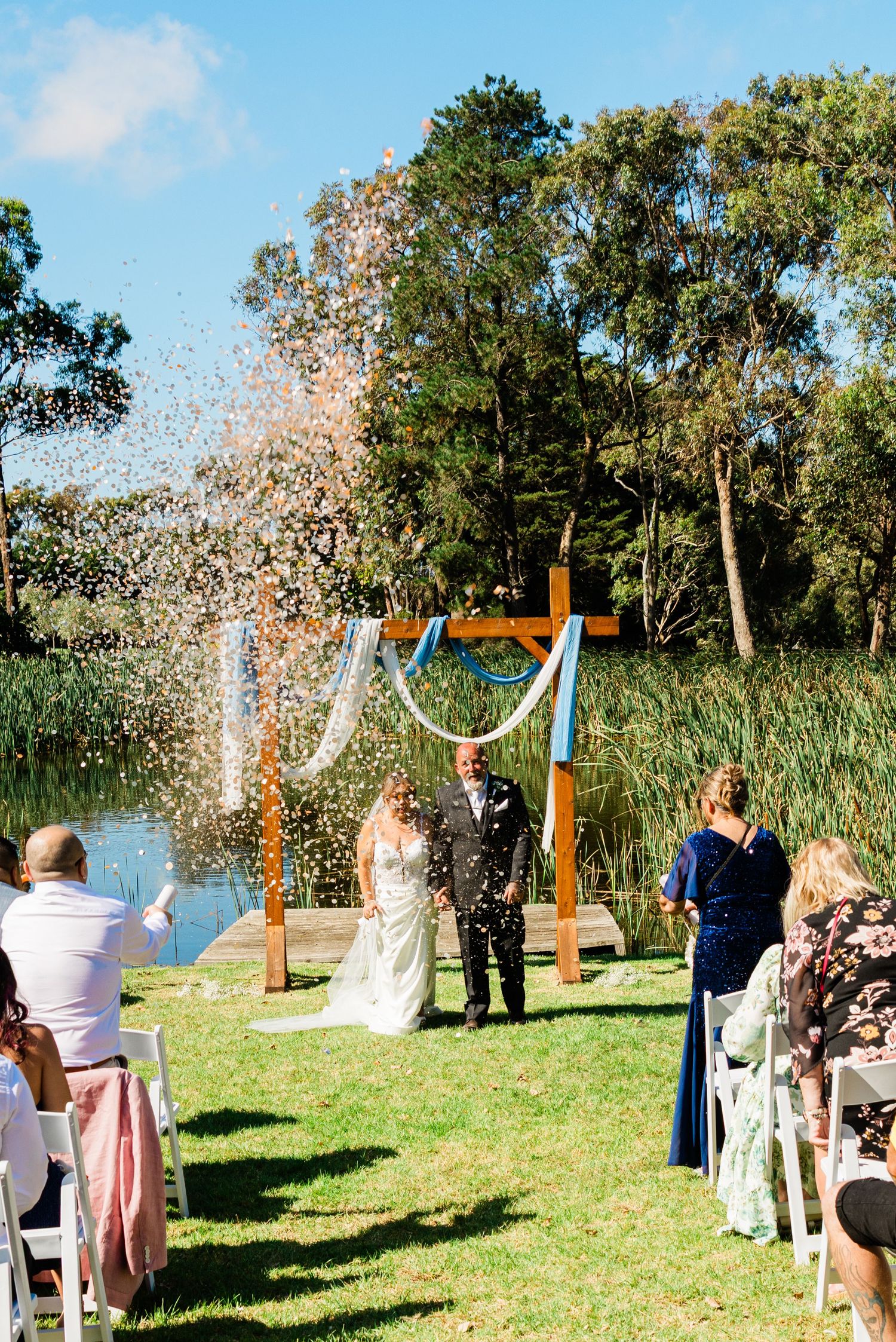 Bride and groom walk down the aisle at outdoor wedding ceremony by a pond, with confetti flying and a wooden arch backdrop.