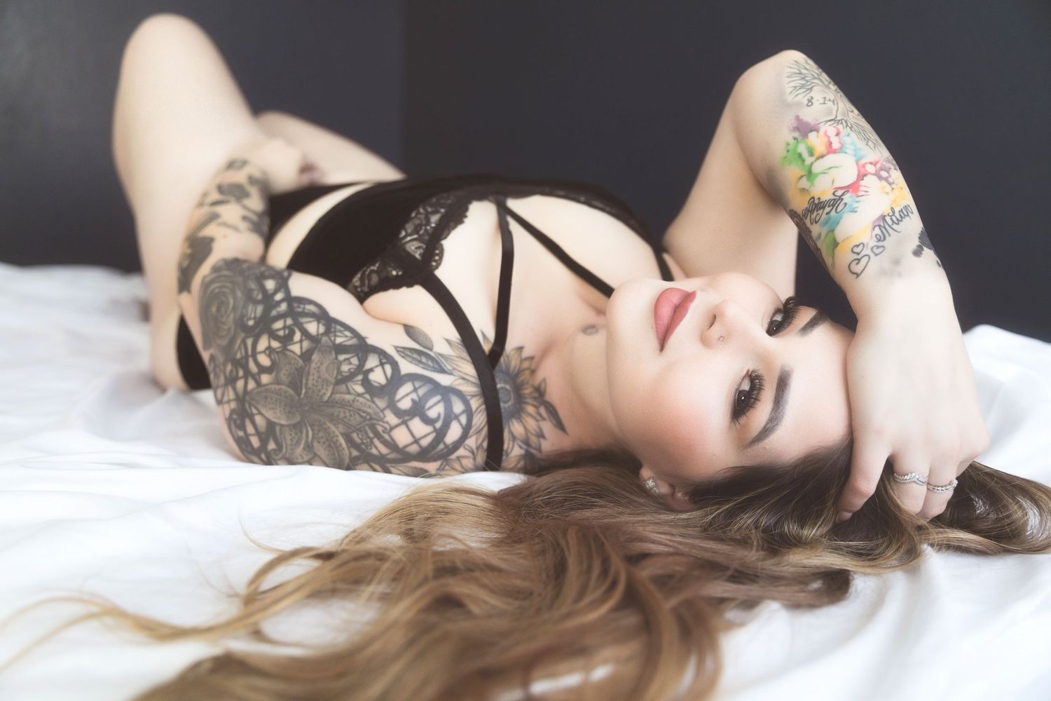 Woman with tattoos lying on white bed wearing black lace lingerie, with long auburn hair and bold makeup.