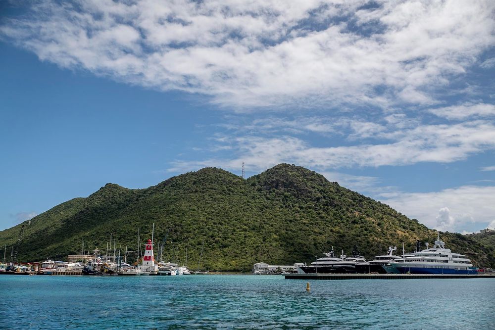Luxury yachts and a red lighthouse dock beside a lush green mountain in a calm Caribbean harbor.