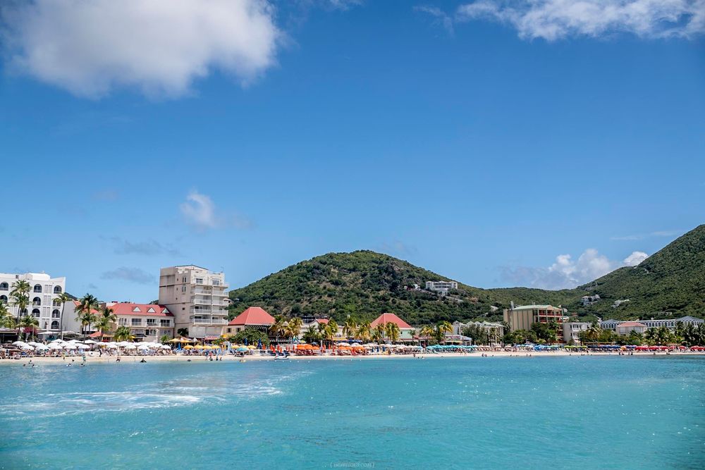Turquoise Caribbean waters front a colorful beach resort town with lush green hills under a bright blue sky.