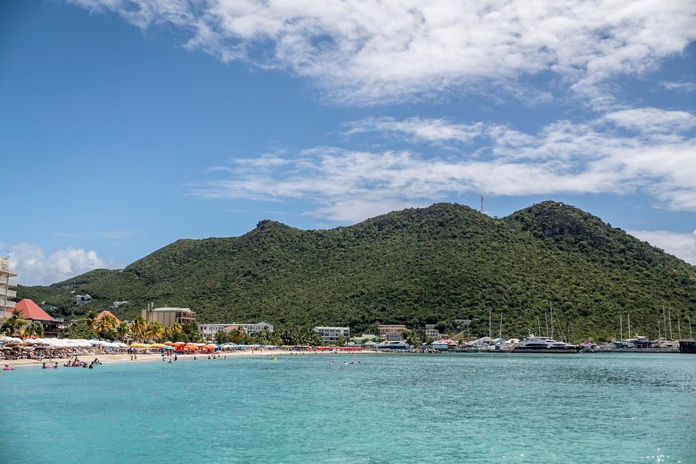 Turquoise Caribbean waters with a sandy beach, lush green mountain, and marina under a bright blue sky in St. Maarten.