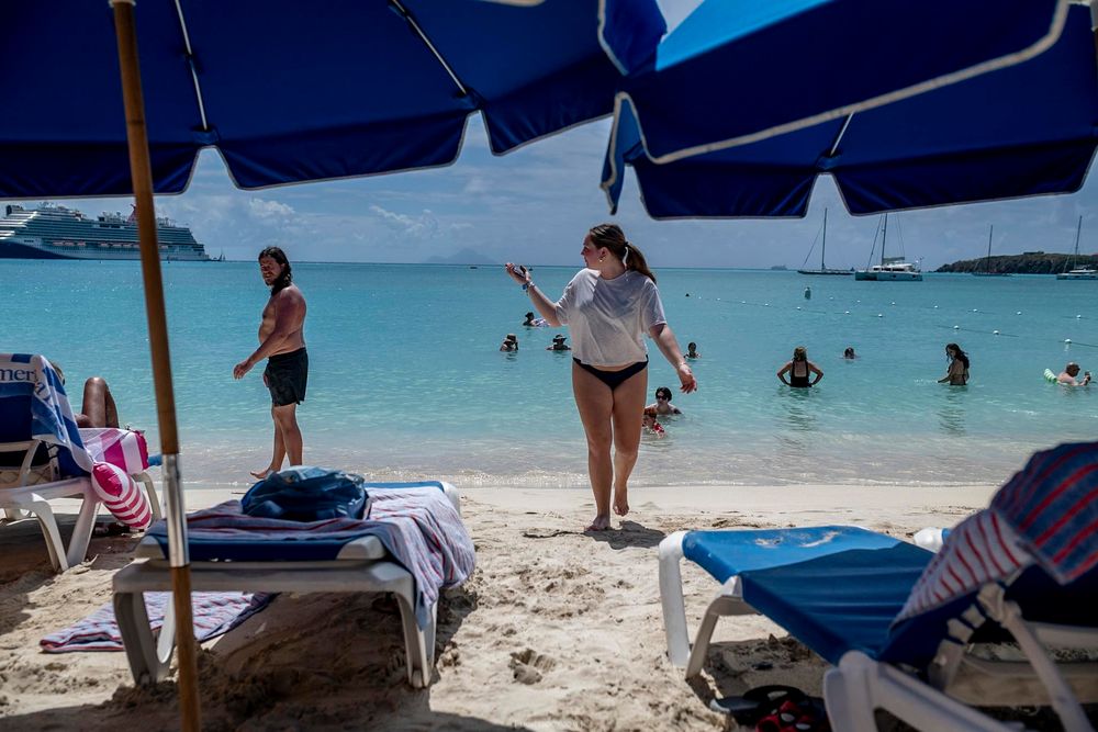 Tourists relaxing under blue beach umbrellas on a sunny Caribbean beach with turquoise water.