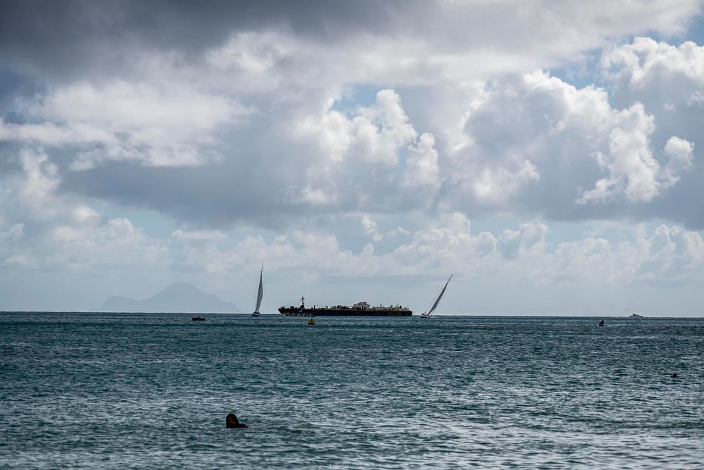 Sailboats and a cargo ship on calm turquoise ocean waters under dramatic cloudy skies, viewed from a tropical beach shore.
