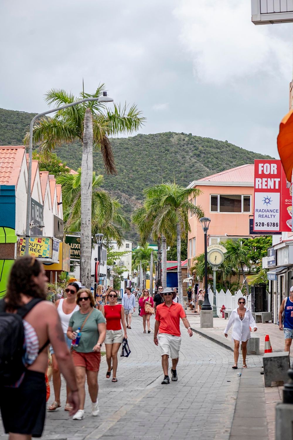 Busy pedestrian shopping street with palm trees, colorful buildings, and a mountain backdrop.