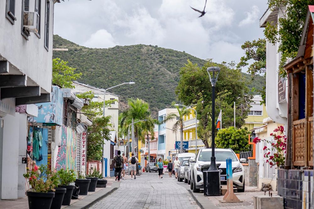 A colorful Caribbean street scene with shops, parked cars, and lush green mountains in the background.