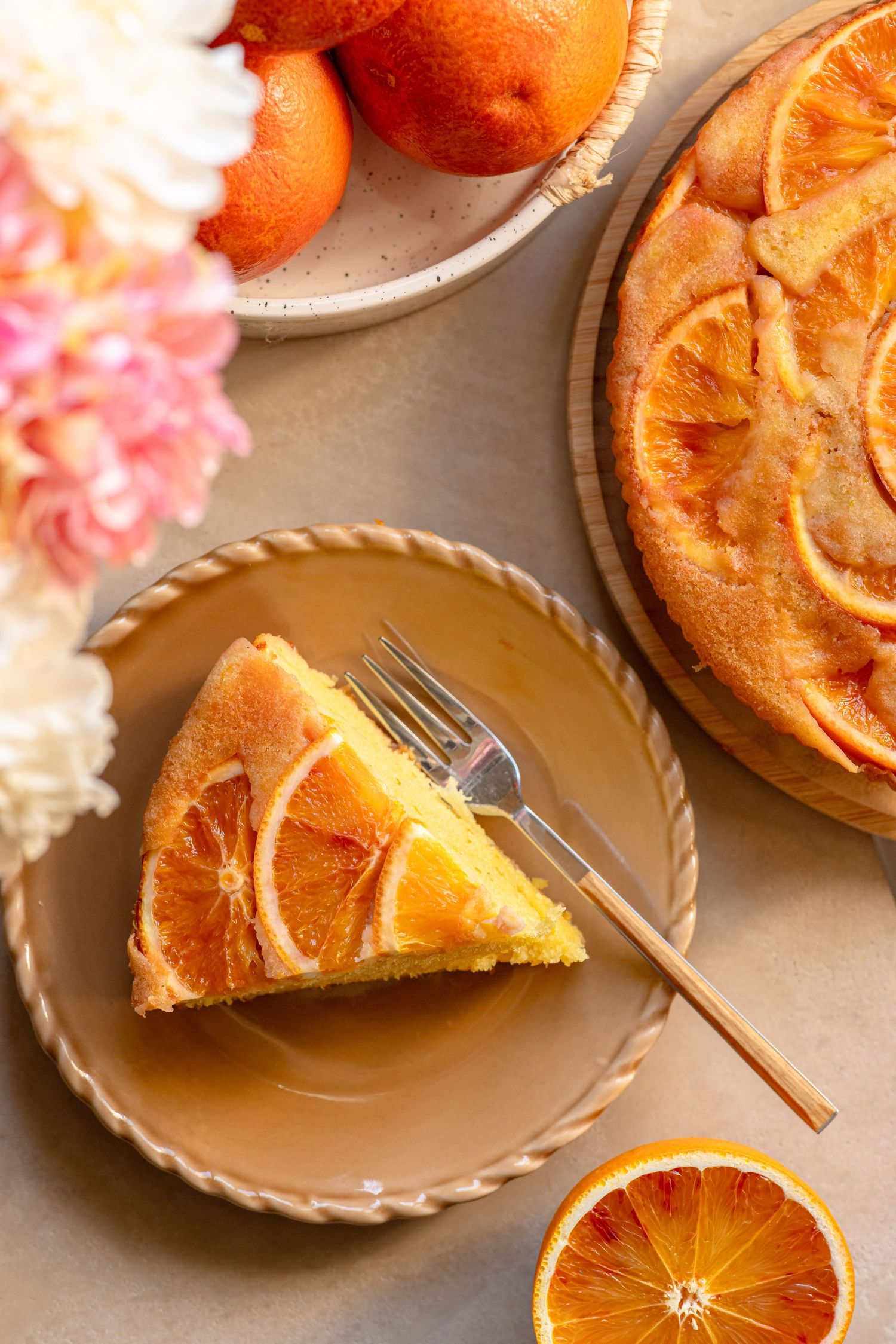 Slice of blood orange upside-down cake on a beige plate with a fork, beside the whole cake and fresh oranges.