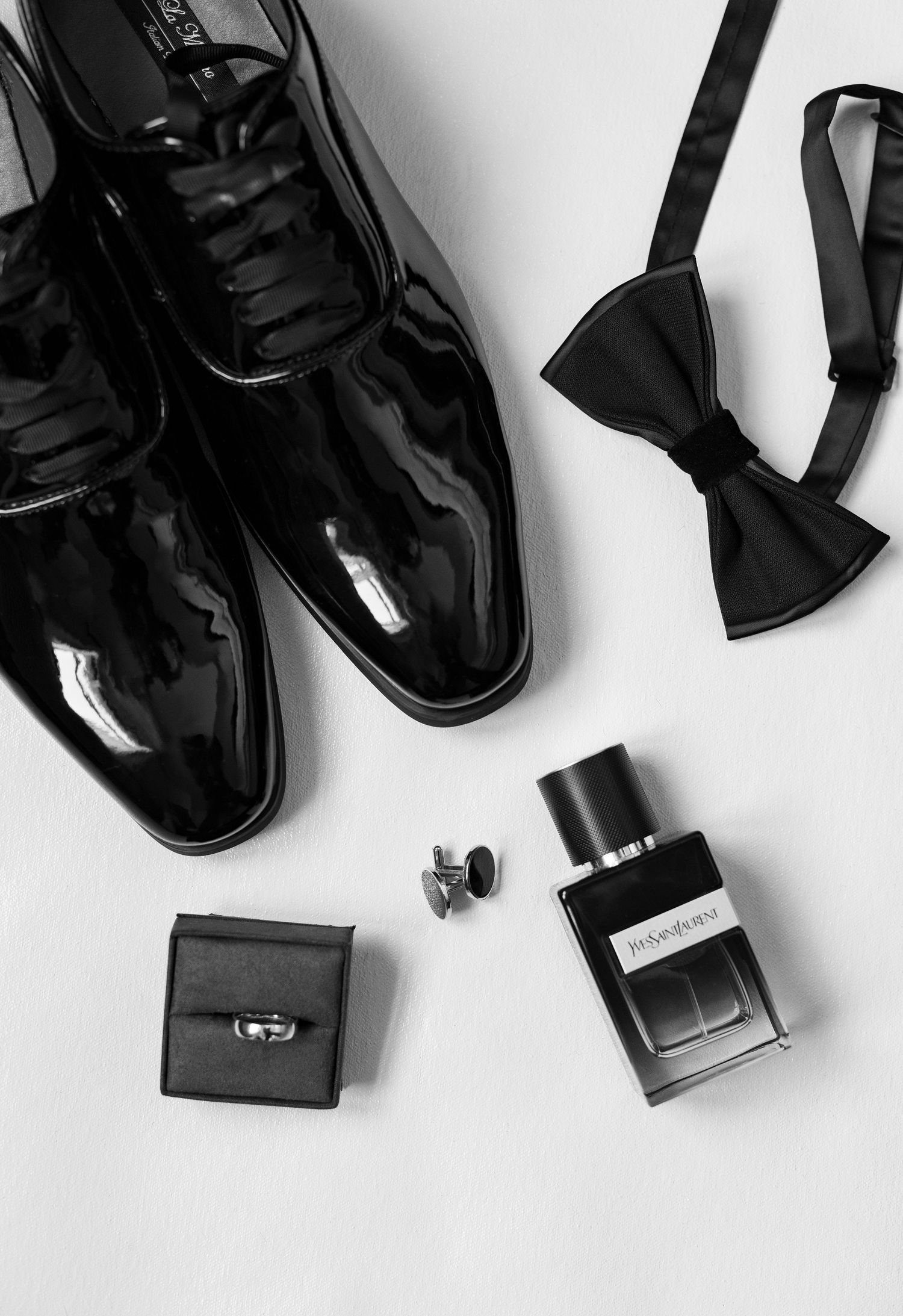 Elegant groom wedding details featuring patent leather tuxedo shoes, black bow tie, wedding band, cufflinks, and cologne during getting read