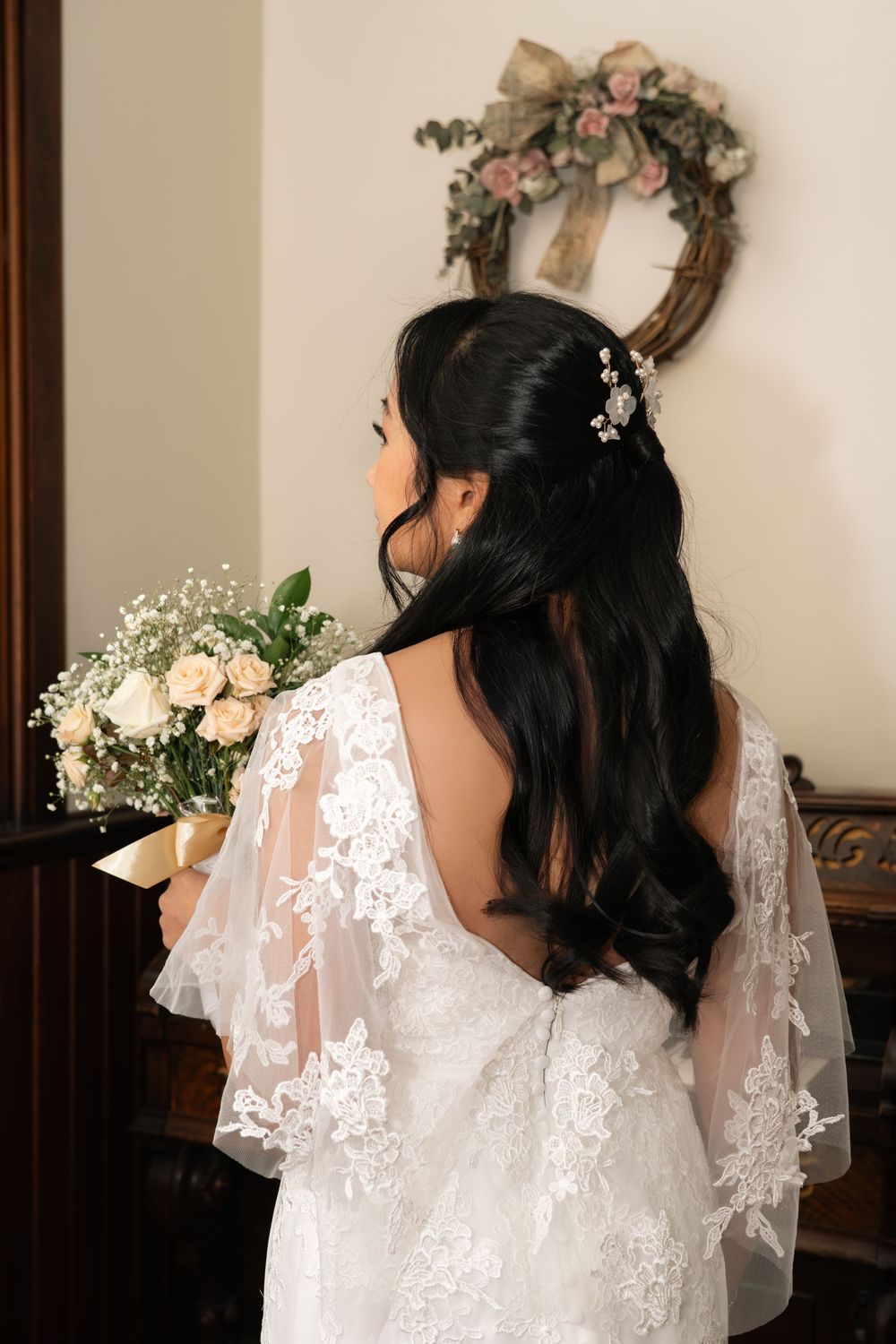 Bride in lace wedding dress with floral hair accessory holds bouquet of roses, standing before a decorative wreath.