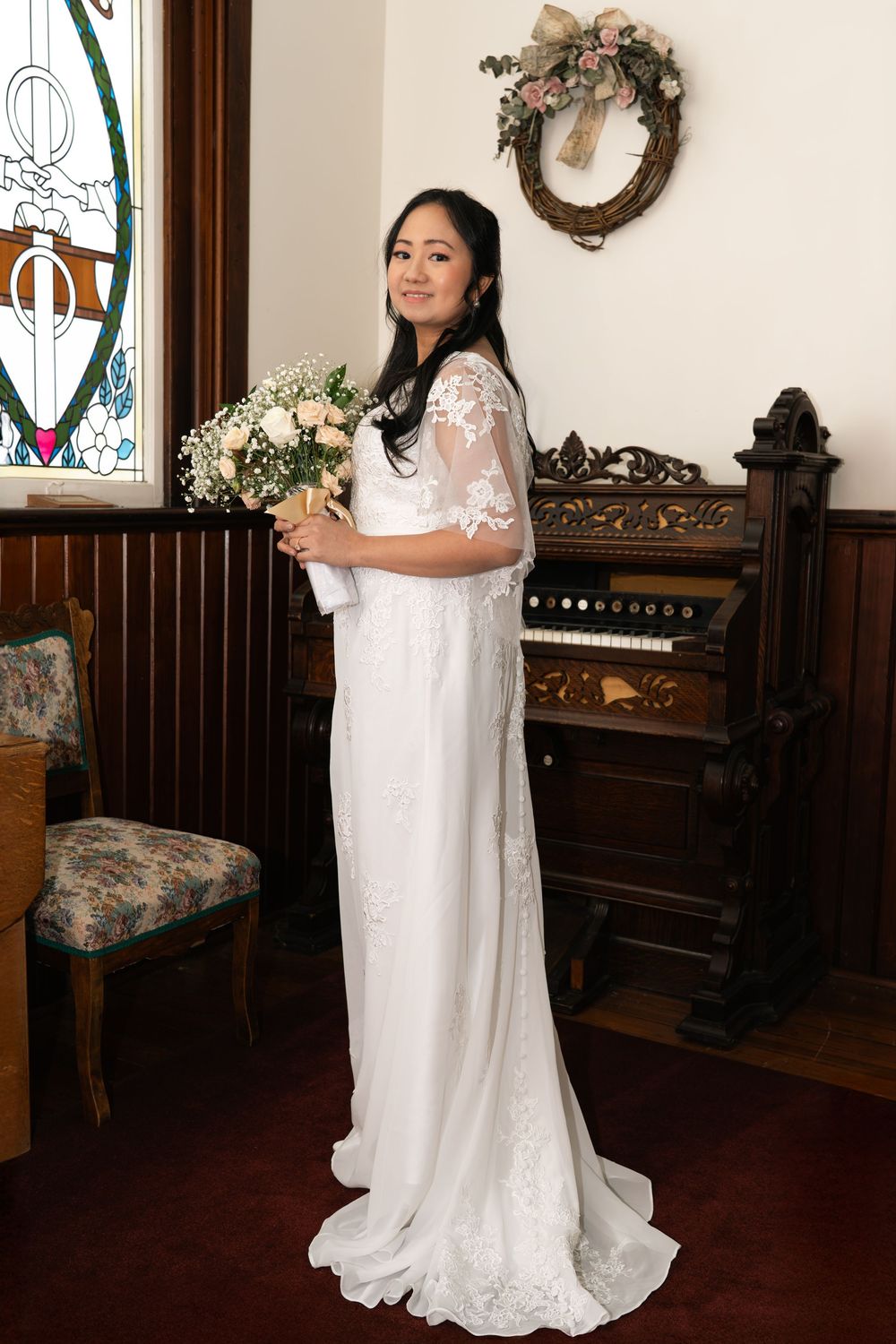 Bride in white lace gown holding bouquet stands by stained glass window and vintage piano.