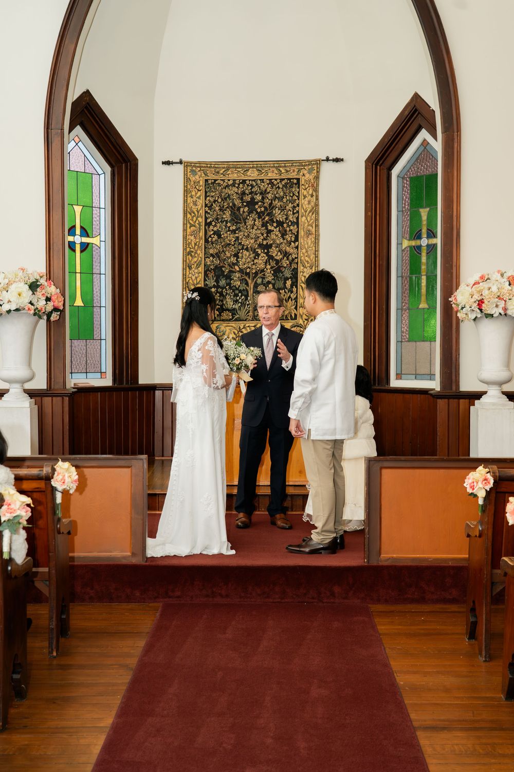 Couple exchanging vows at chapel altar with officiant, stained glass windows, and floral arrangements on wooden pews.