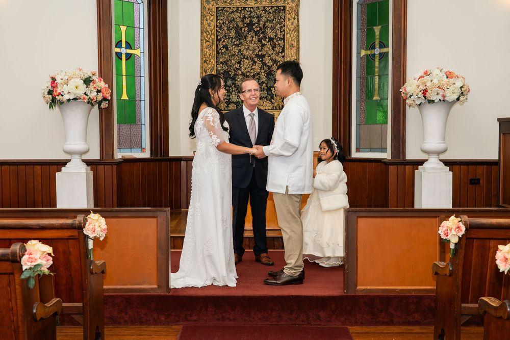 Couple exchanges vows at church wedding altar with officiant, floral arrangements, and guests in elegant chapel setting.