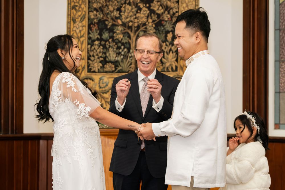 Couple holding hands during intimate wedding ceremony with officiant smiling, small child in white dress nearby.