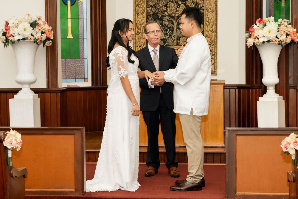 Couple exchanging vows at church altar with officiant, bride in white gown, groom in white shirt, floral arrangements.