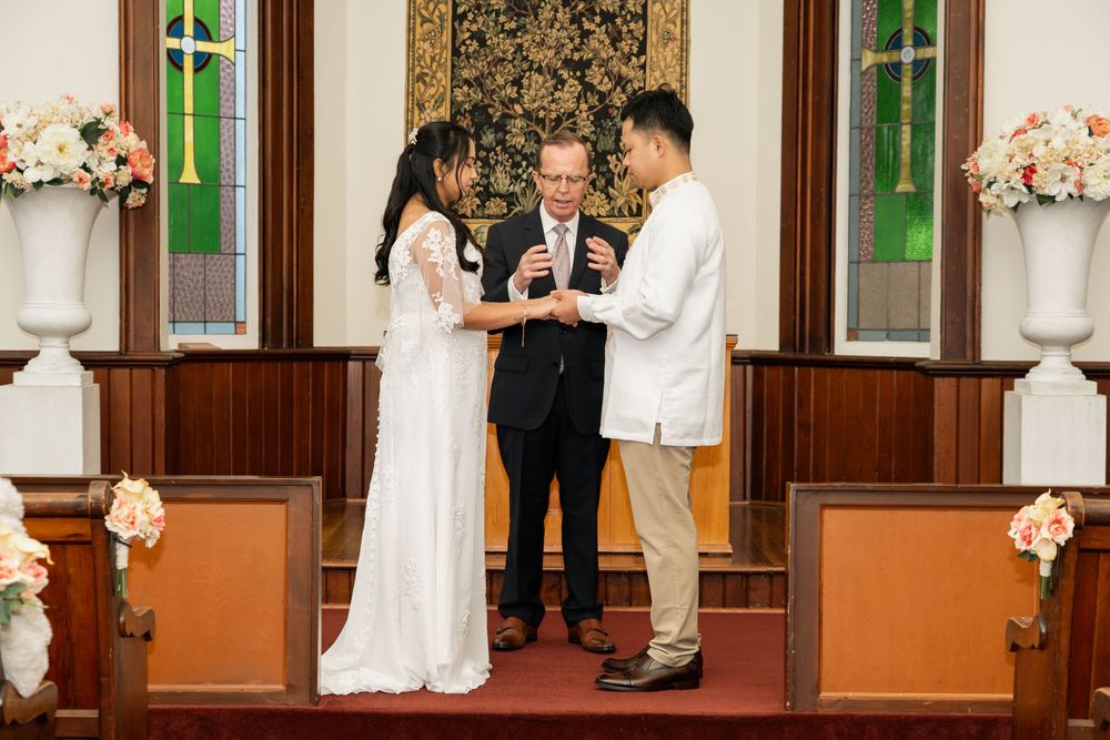Couple exchanging wedding vows at church altar with officiant, floral arrangements, and stained glass windows in background.