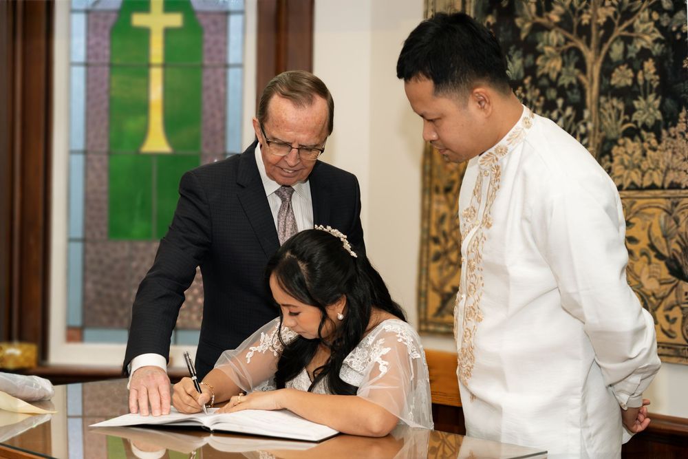 Bride signs wedding registry document with officiant and groom watching in formal church ceremony room.