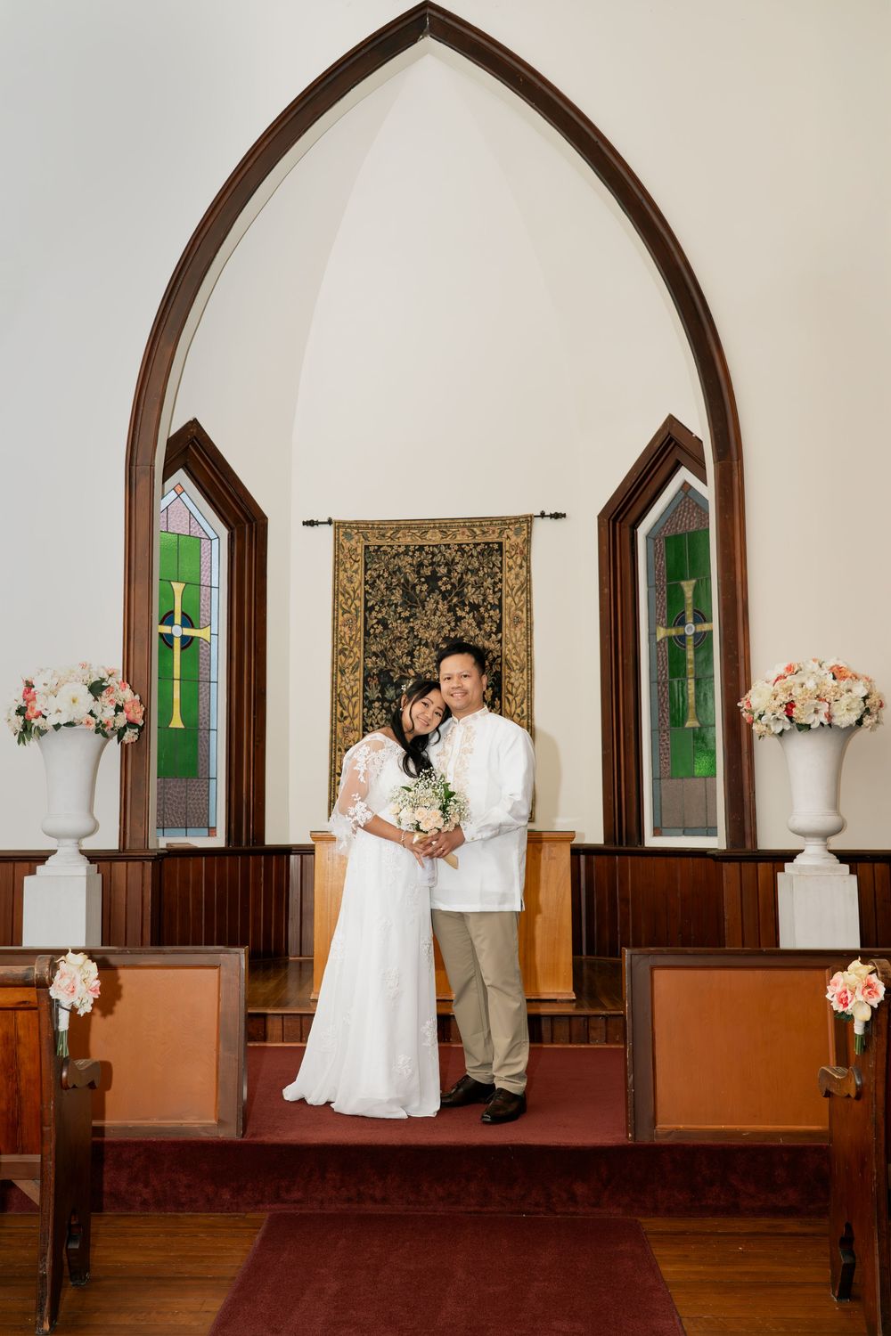 Couple in white wedding attire poses at church altar with stained glass windows and floral arrangements.