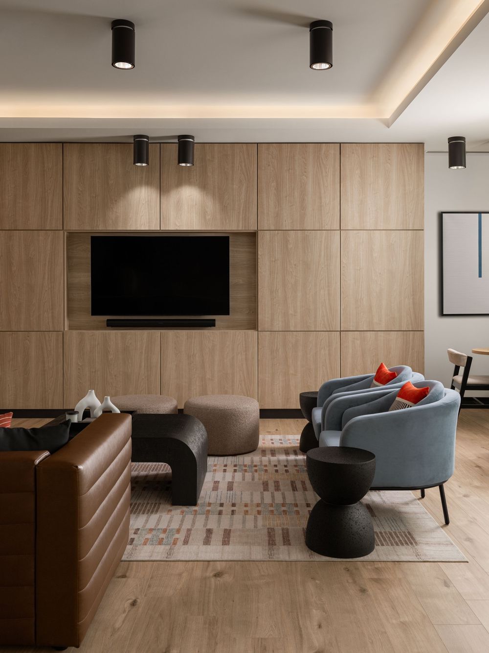 Contemporary living room featuring wood panel wall, mounted TV, blue armchairs, brown leather sofa, and colorful rug.