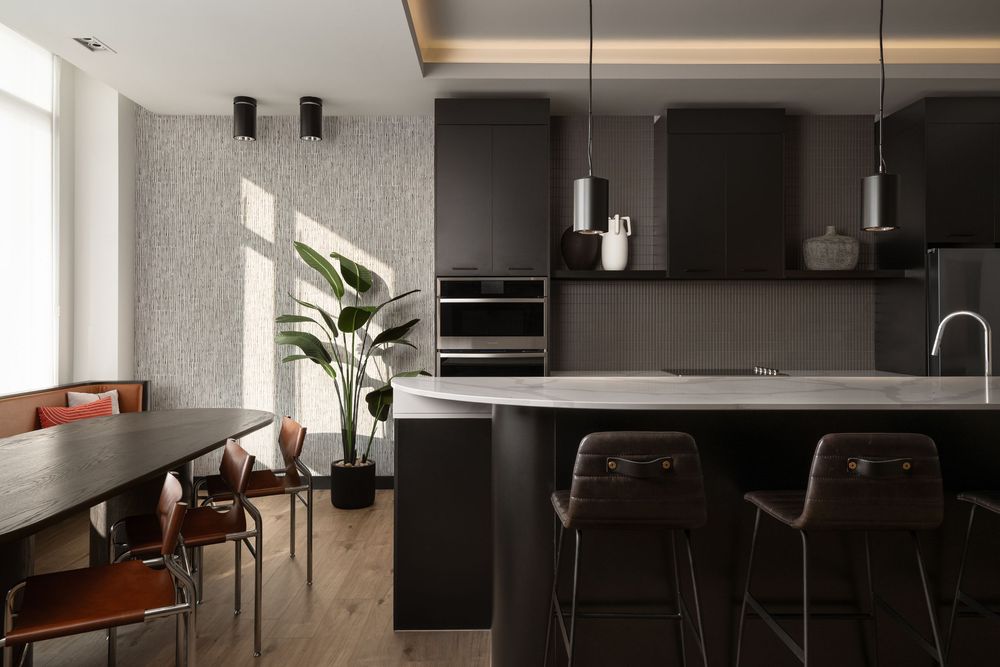 Modern dark kitchen with black cabinetry, marble island, pendant lights, bar stools, and a dining area with leather chairs.