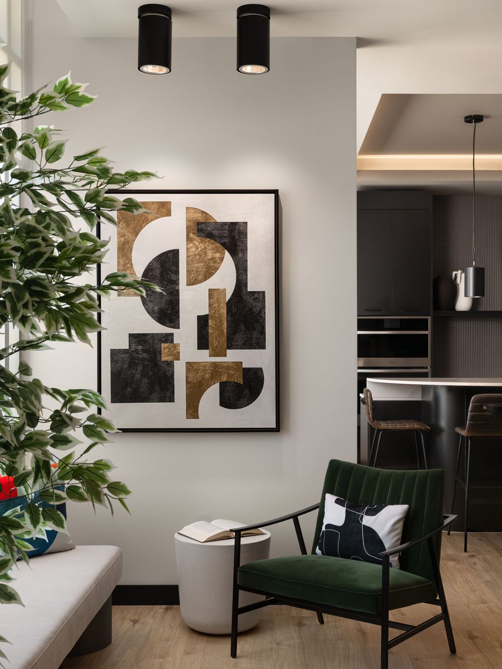Modern living room with green velvet chair, abstract gold and black wall art, indoor plant, and open kitchen in background.