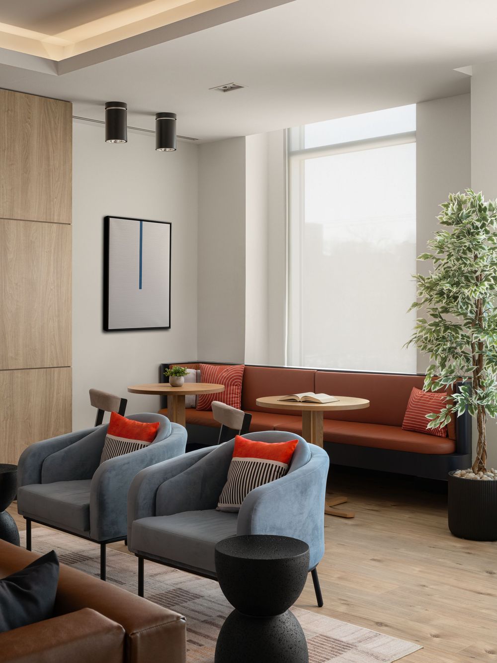 Modern hotel lobby lounge with gray armchairs, red cushions, brown sofa, wood floors, and a large indoor tree by bright windows.