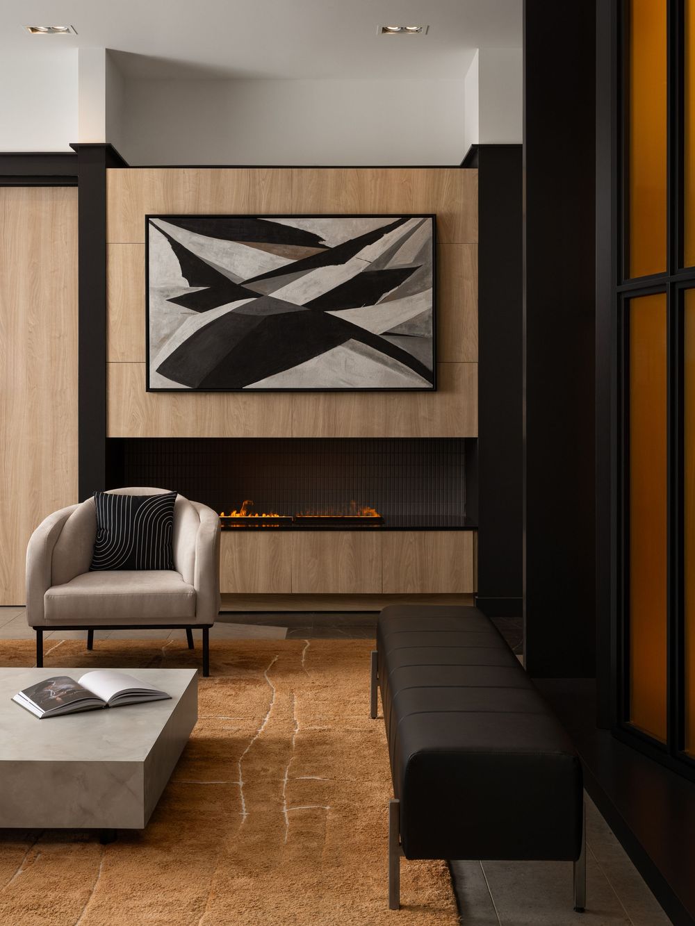 Modern living room with linear fireplace, wood paneling, abstract art, gray armchair, marble coffee table, and black bench seating.