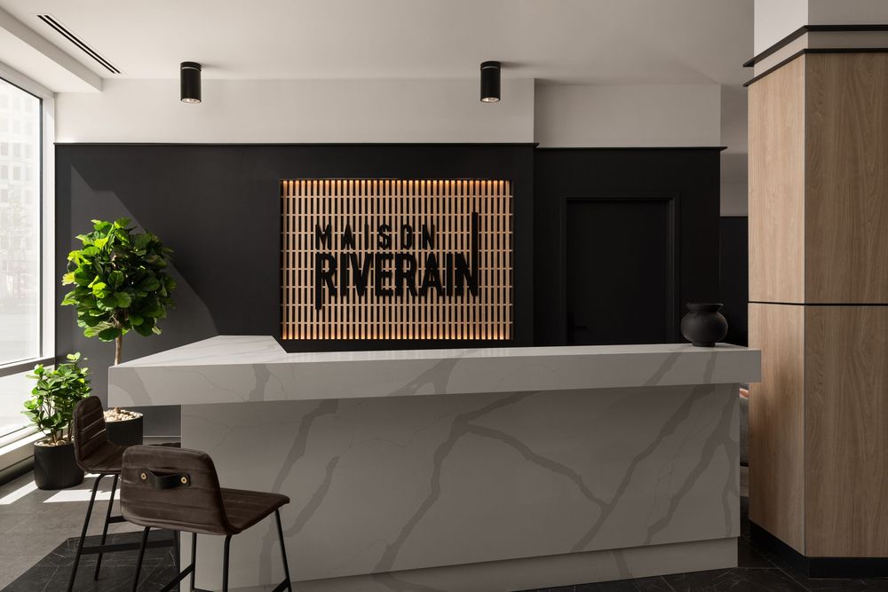Modern hotel reception desk with marble counter and wooden Maison Riverain sign against a dark accent wall.