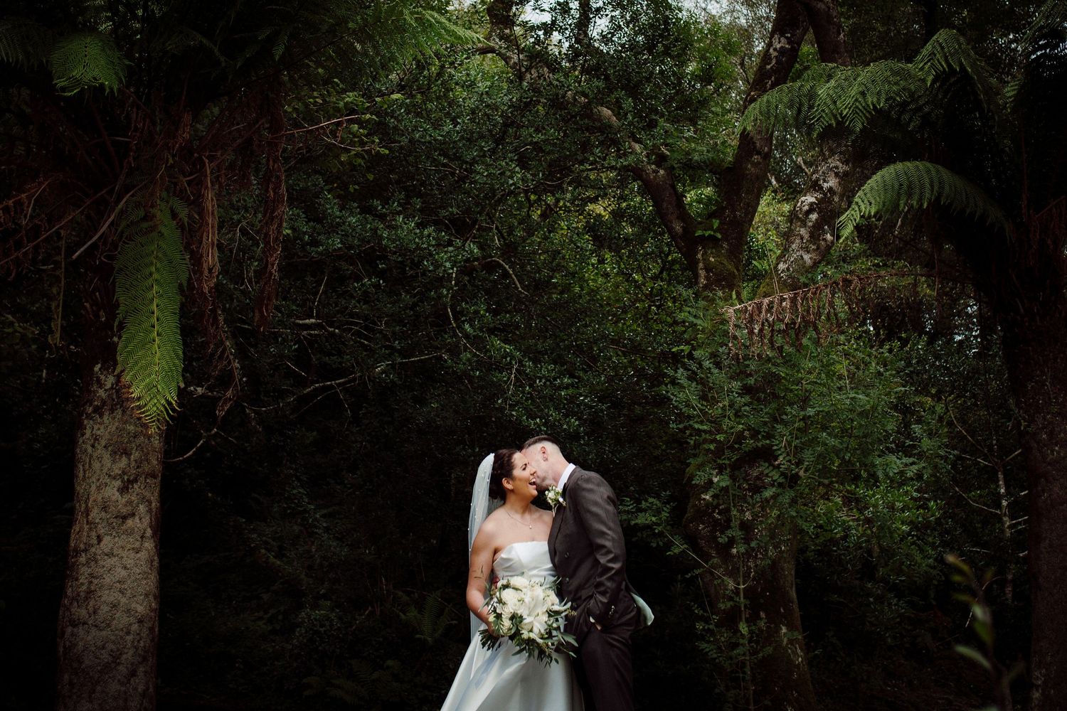 Bride and groom share a romantic kiss surrounded by lush green trees and ferns in a forest wedding setting.
