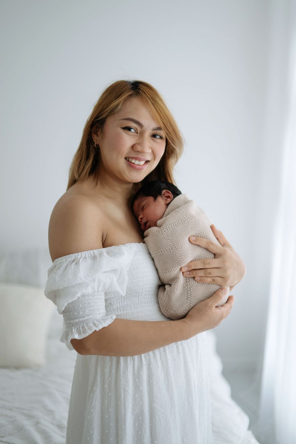 Smiling mother in white off-shoulder dress tenderly holding her swaddled newborn baby against her chest.