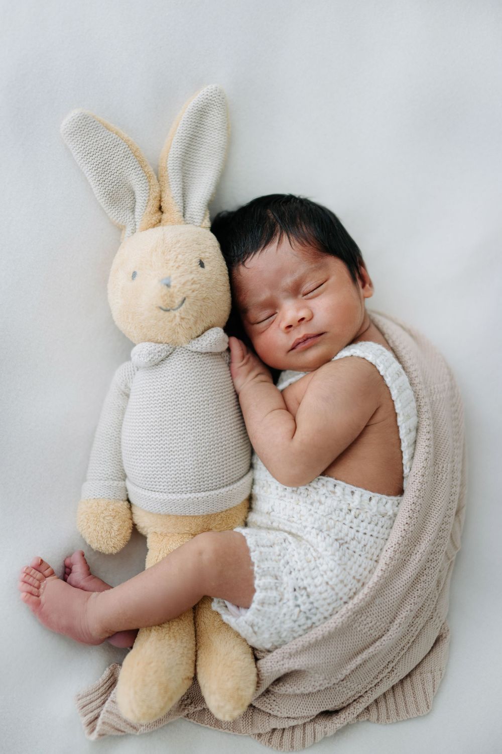 Newborn baby sleeping beside a soft bunny stuffed animal in white knit outfit.