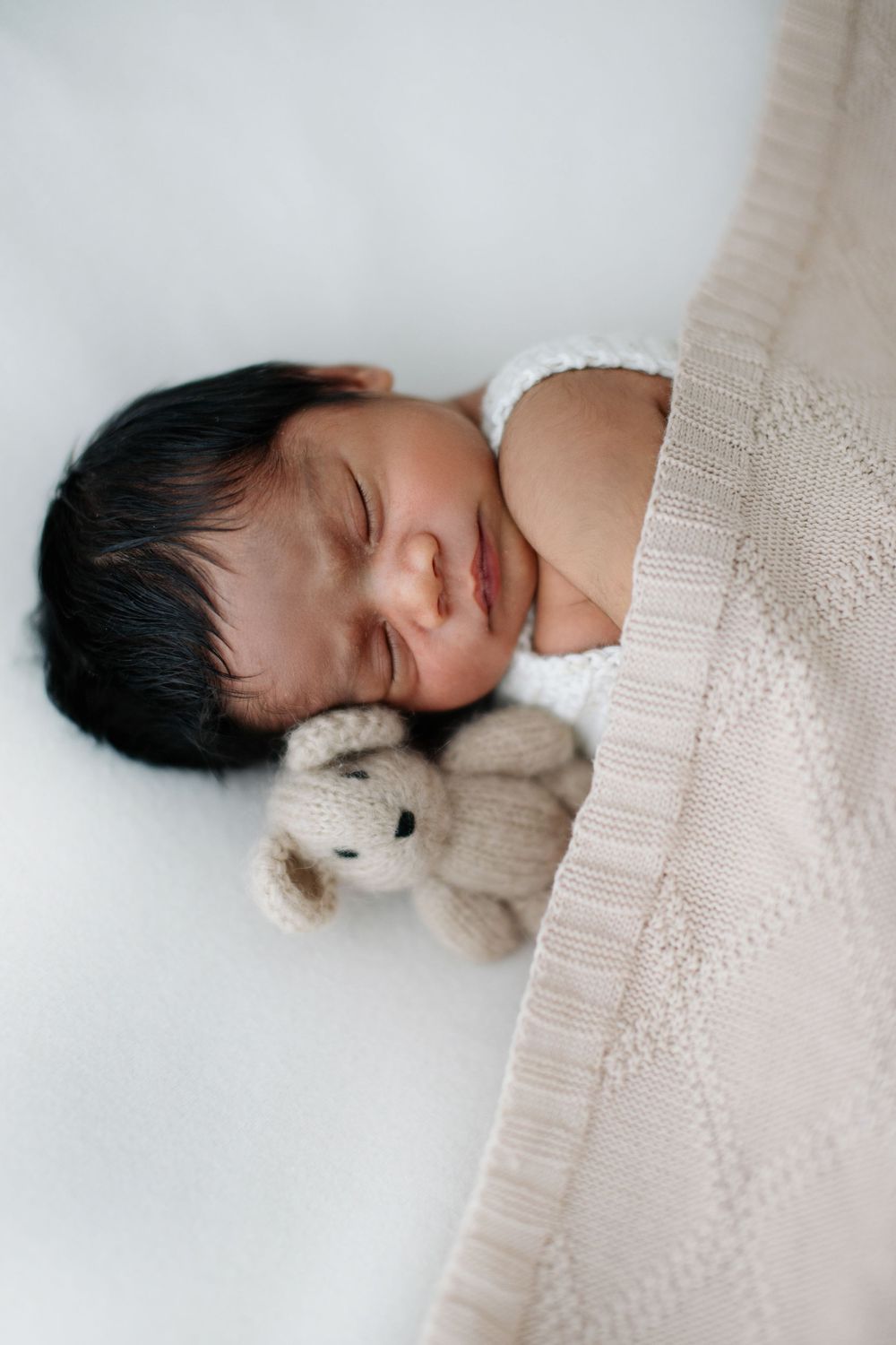 Sleeping newborn baby with tiny teddy bear tucked under beige knit blanket.