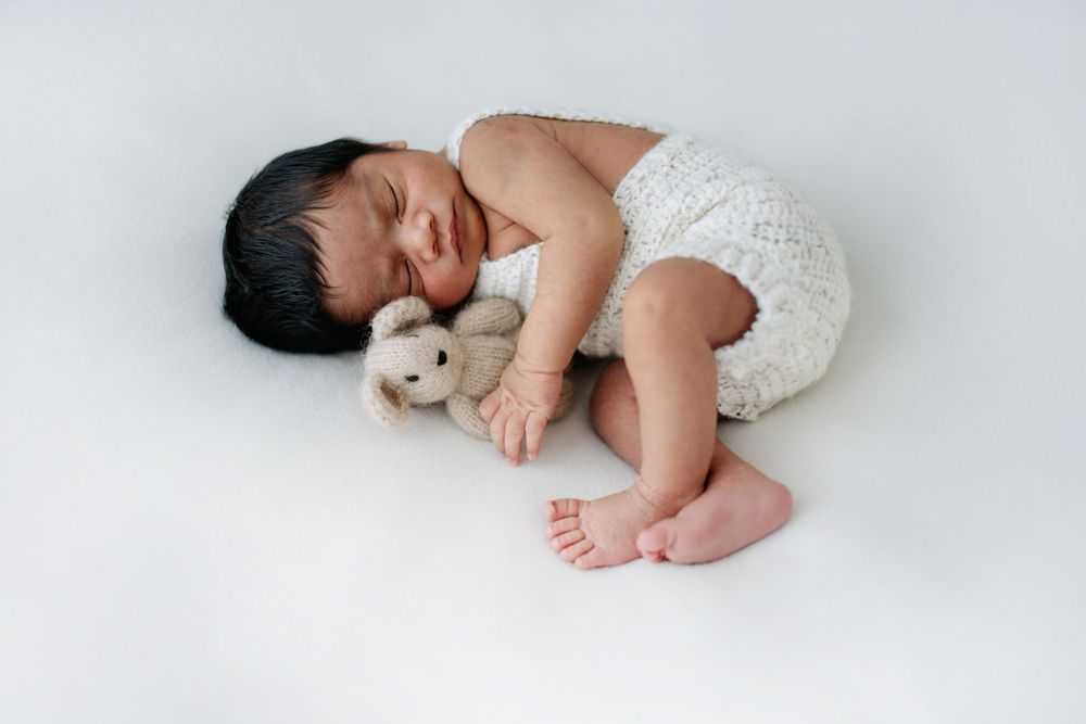 Newborn baby sleeping curled up with small teddy bear on white background.