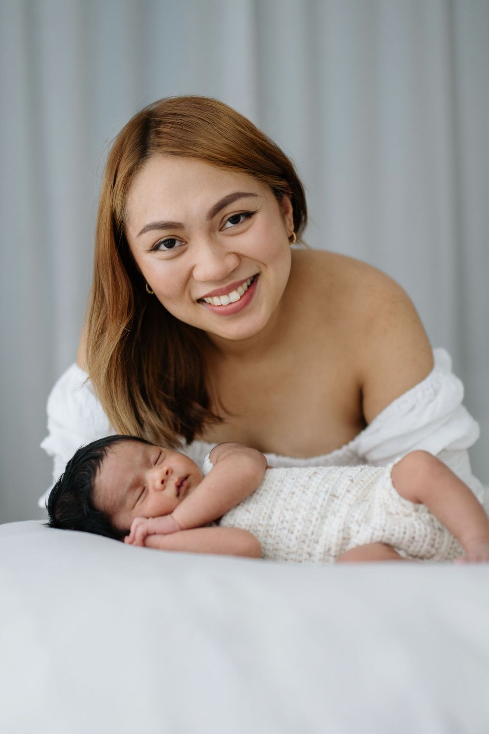 Smiling young mother posing with her sleeping newborn baby wrapped in a white knit blanket on a bed.