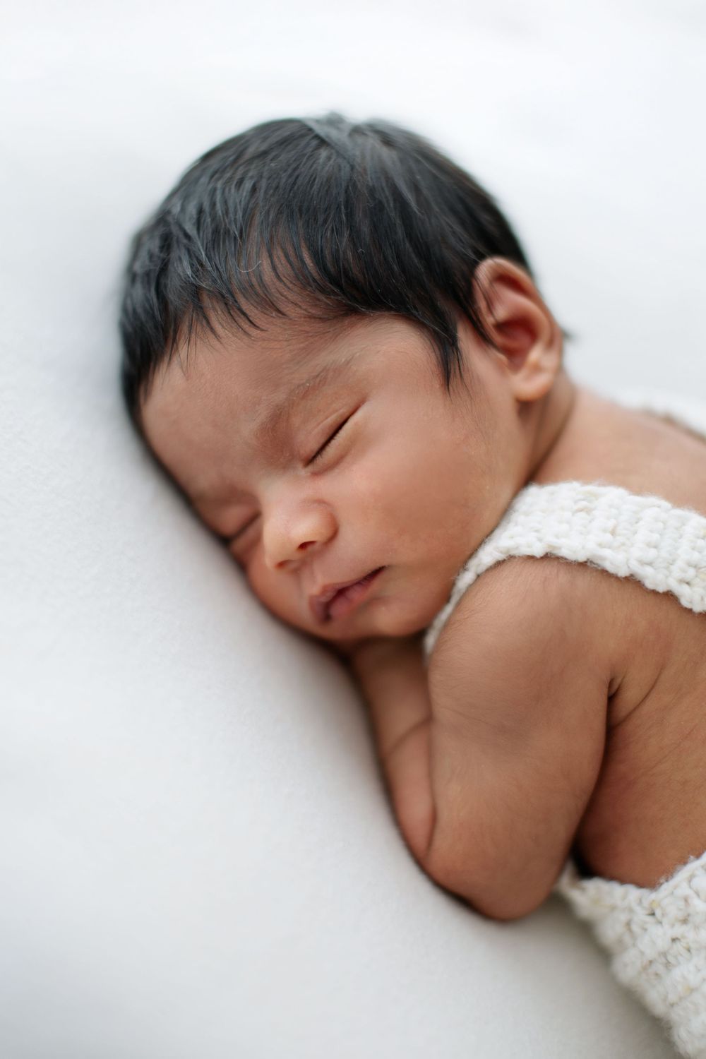 A peaceful newborn baby with dark hair sleeps on a white blanket wearing a delicate white knit outfit.
