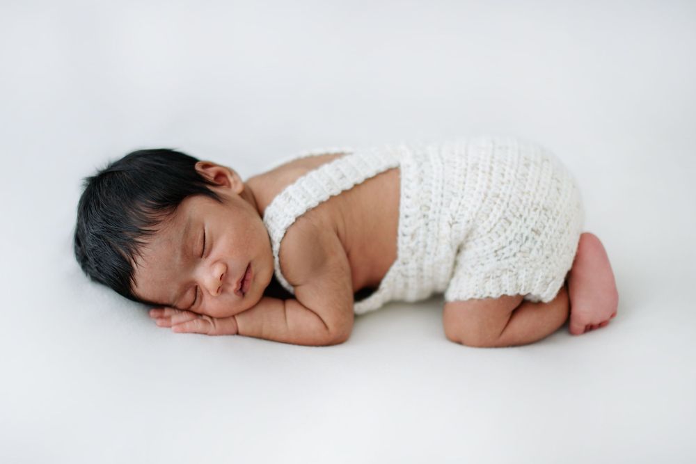 Sleeping newborn baby in white knit romper posed curled up on white background.