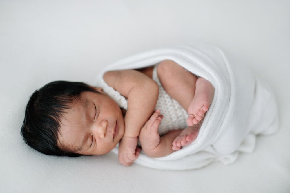 Peaceful newborn baby with dark hair sleeping curled up in a white knit wrap on a soft white background.