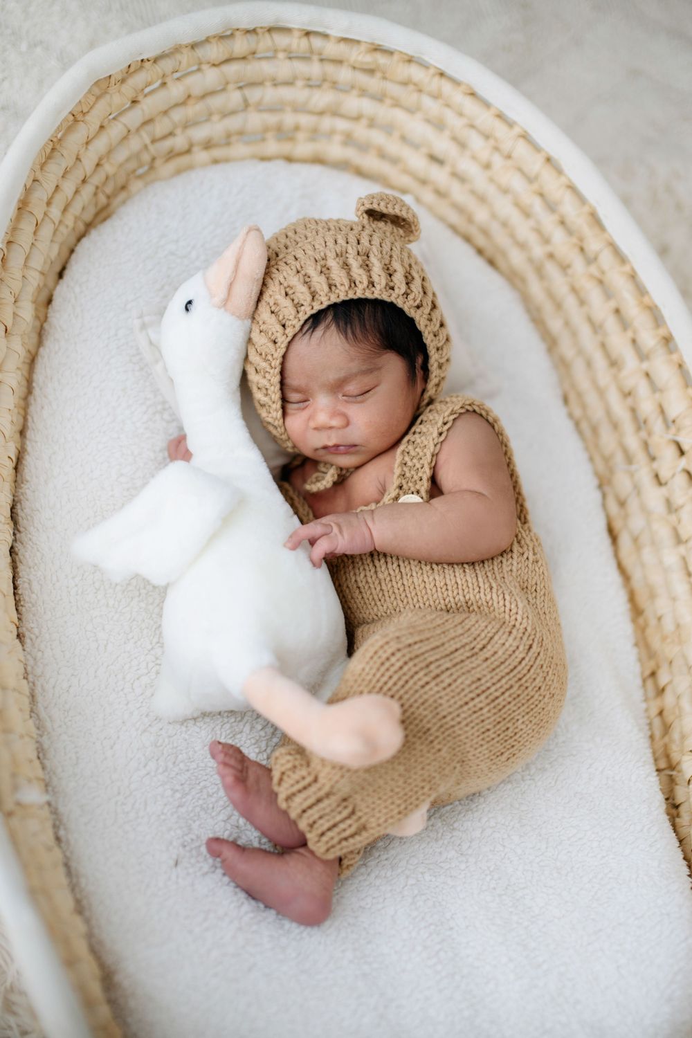 Newborn baby sleeping in a wicker Moses basket wearing a knit bear romper and bonnet, cuddling a white bunny stuffed animal.