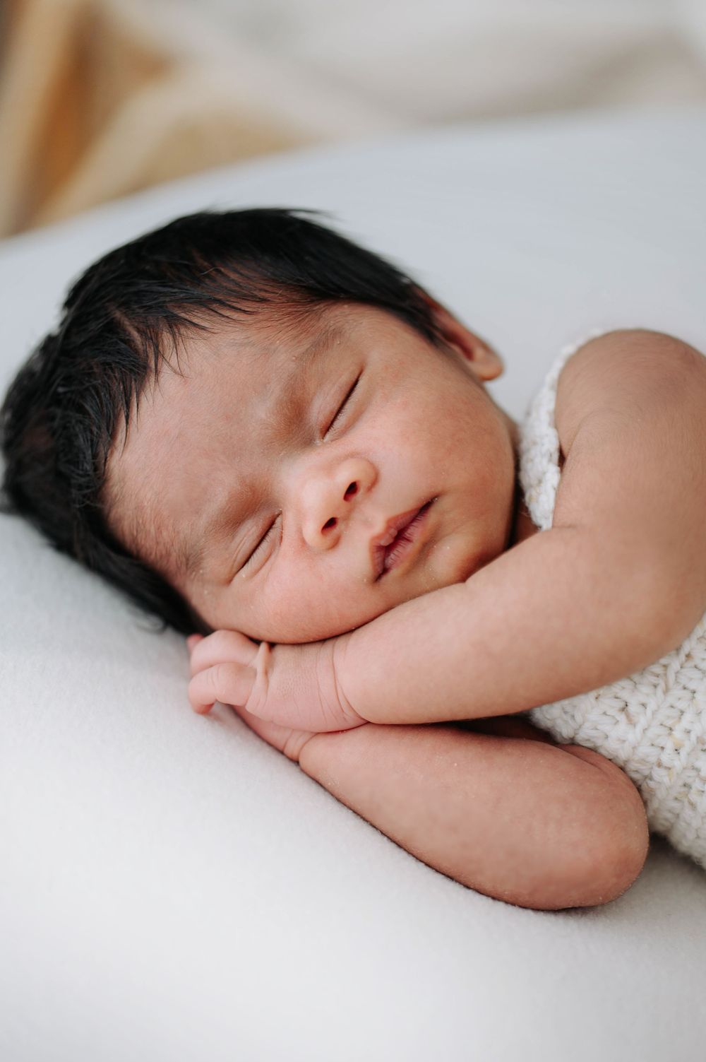 Peaceful newborn baby with dark hair sleeping soundly, hands tucked under cheek, wrapped in white knit blanket.