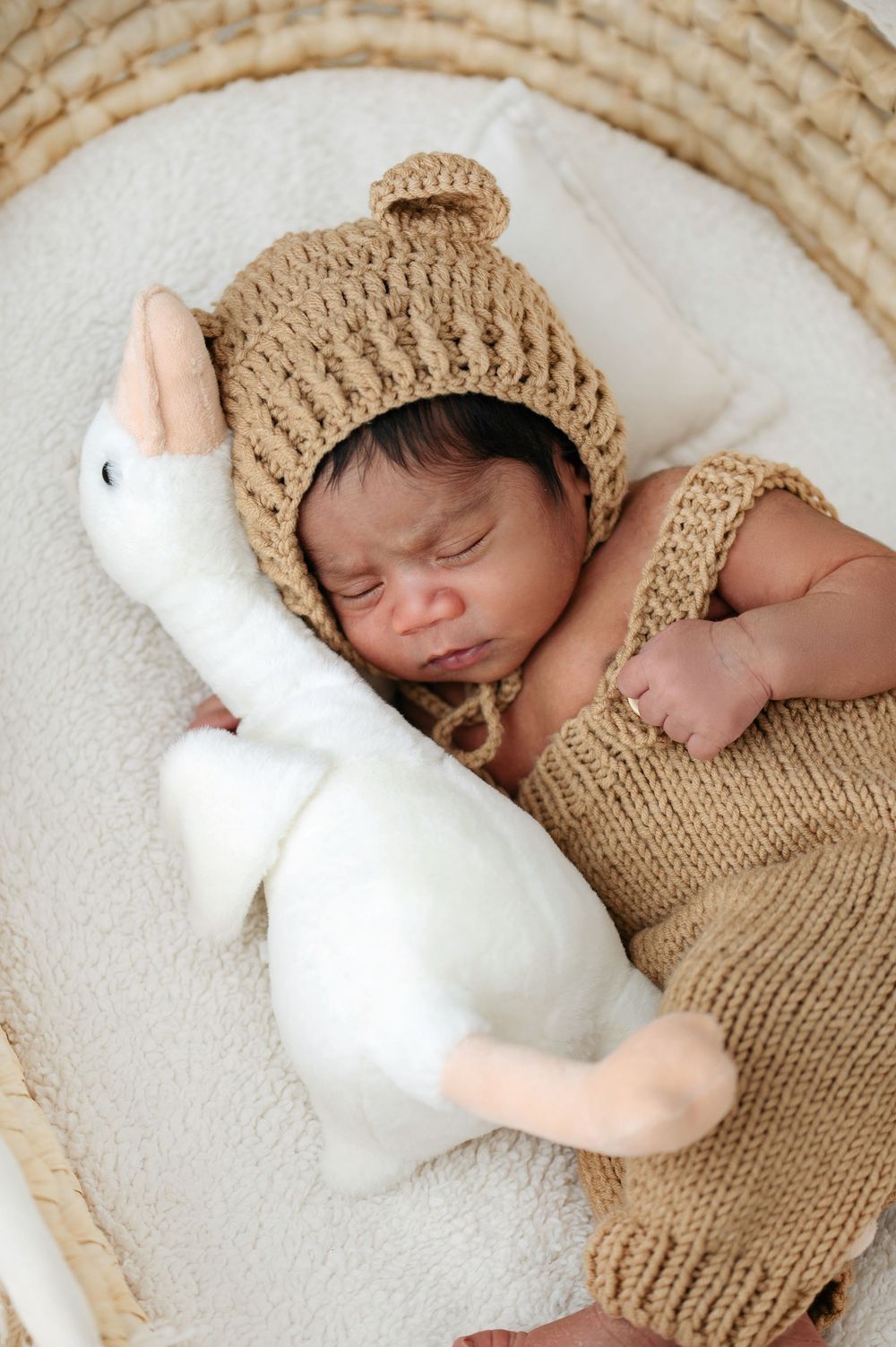 Sleeping newborn baby in knit bear hat and romper cuddling a white duck stuffed animal in a wicker basket.