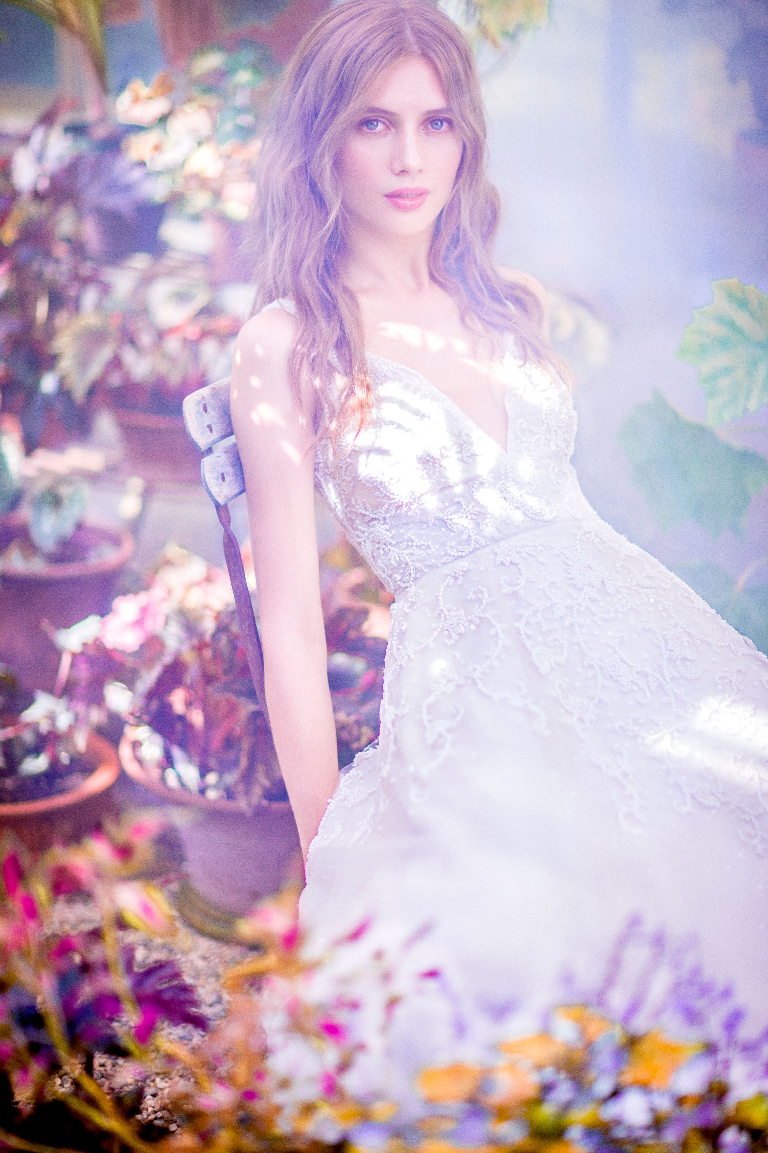 Woman in elegant white lace wedding gown surrounded by colorful flowers in a dreamy, misty garden setting.