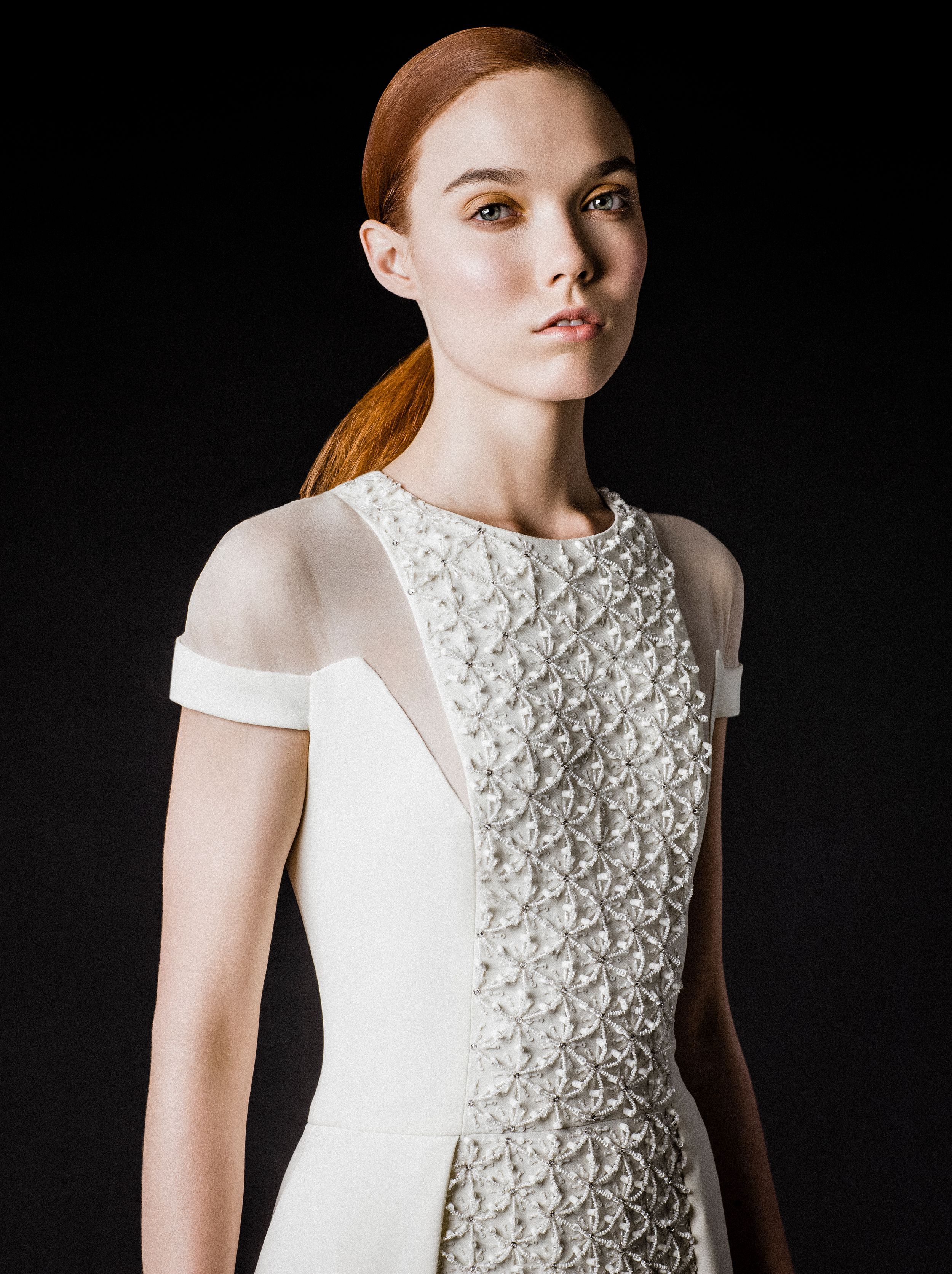 Model wearing elegant white textured floral embroidered dress with sheer cap sleeves against dark background.