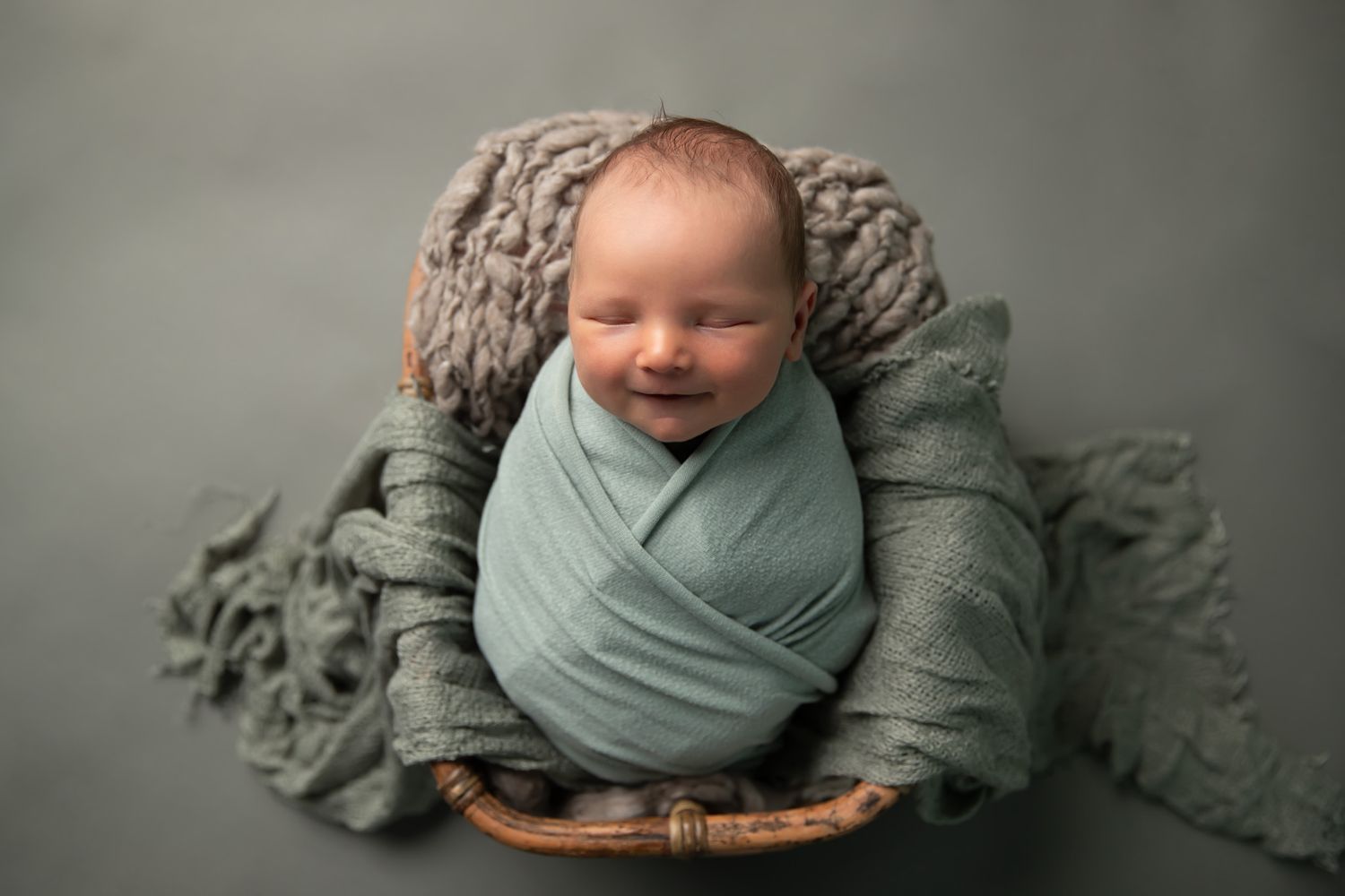Peaceful newborn baby swaddled in teal wrap smiling in wicker basket on gray studio background.
