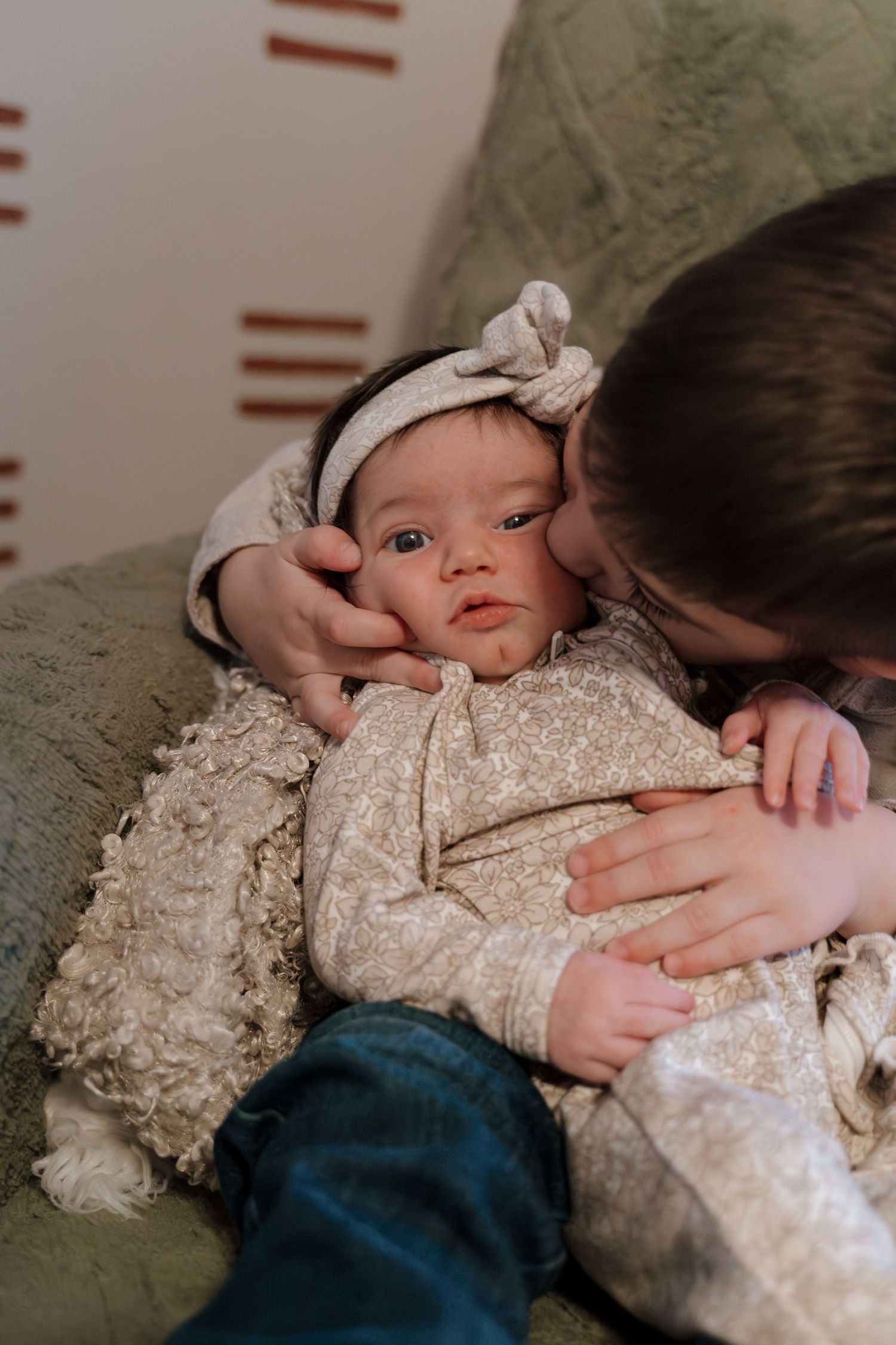 Older child gently kissing a newborn baby wearing a white bow headband and cream outfit on a green sofa.