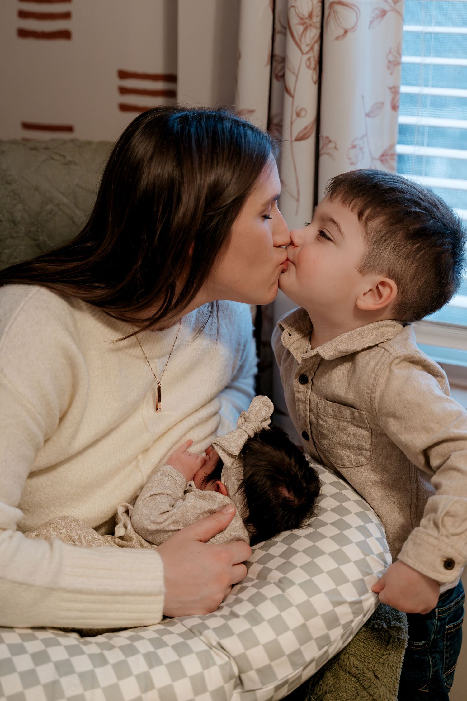 A mother kisses her young son while cradling a newborn baby on a nursing pillow indoors.