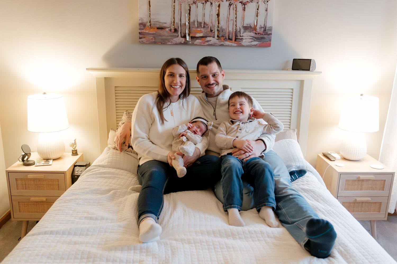 Happy family of four sitting on a bed, parents smiling with toddler and newborn baby in cozy bedroom.