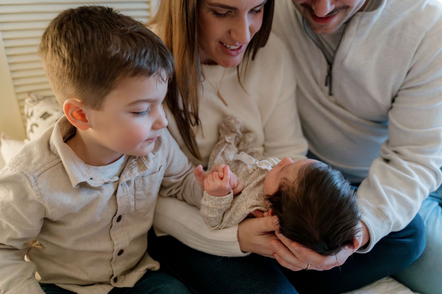 Family of four admiring newborn baby, parents and young boy gathered closely together in warm indoor setting.